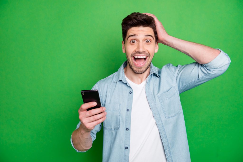 Photo of cheerful brunet haired excited crazy casual man holding telephone, rejoicing with feednews read just shouting with ecstatic emotions isolated over green vivid color background